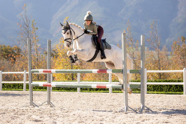 Equestrian rider jumping a grey horse in LeMieux AW25 Rosemary base layer and Damson riding leggings. Outdoor autumn arena setting, showcasing LeMieux AW25 colour combination for horse and rider.